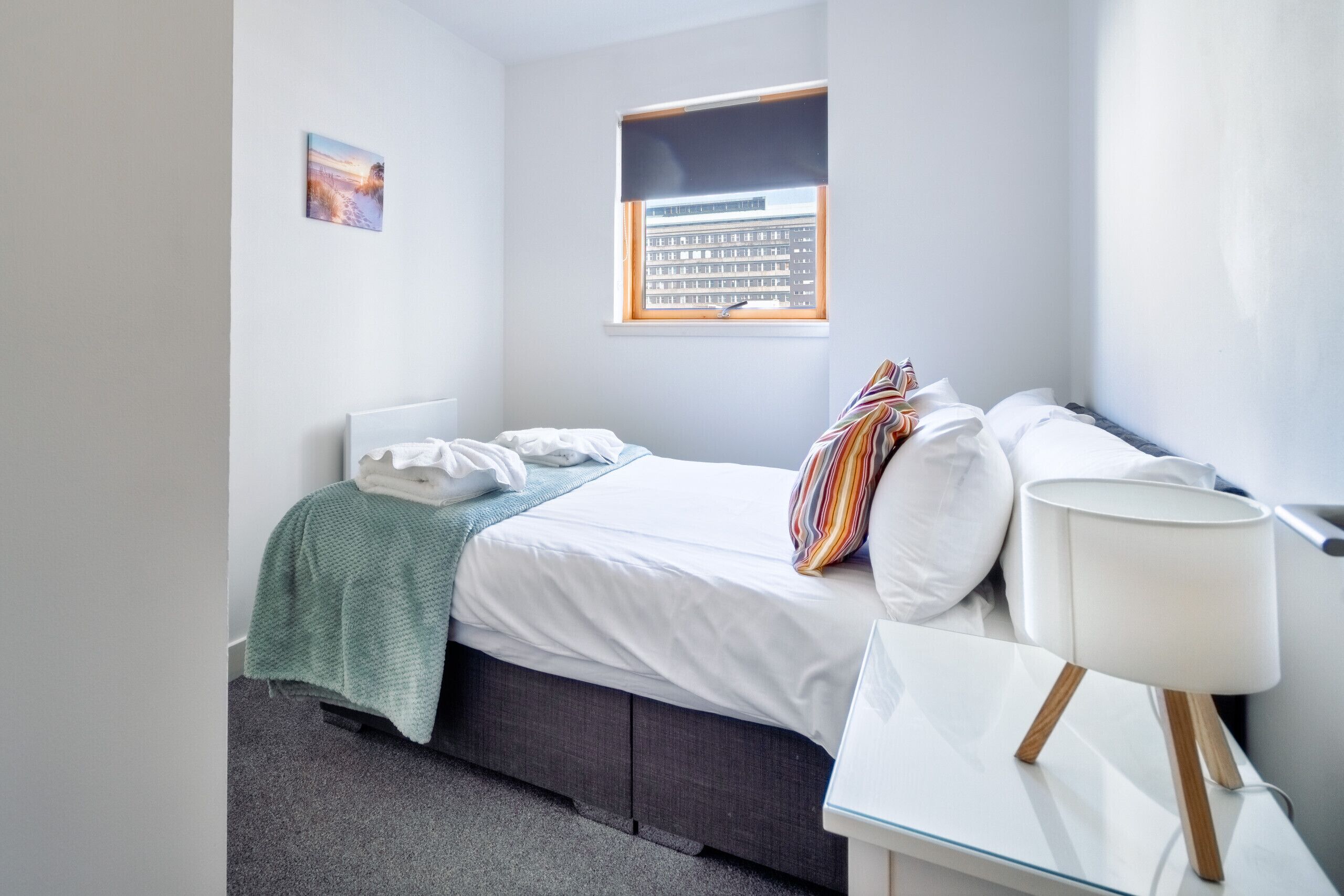 Een keurig opgemaakt slaapkamer met een bed, een bedlampje, een raam met jaloezieën en een klein schilderijtje aan de muur.