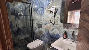 Et badeværelse med marmorvæg, brusekabine i glas, toilet, vask med spejl og en hårhætter fastgjort på væggen.