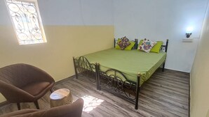 A small, single-bed room with a green bedspread, a brown chair, and a small round table. - Hype Hostel Udaipur (Udaipur)