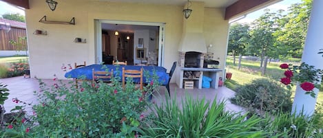 Un patio cubierto con una mesa, sillas, una parrilla y un jardín con flores y plantas.