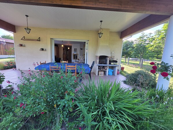 Un patio cubierto con una mesa, sillas, una parrilla y un jardín con flores y plantas.