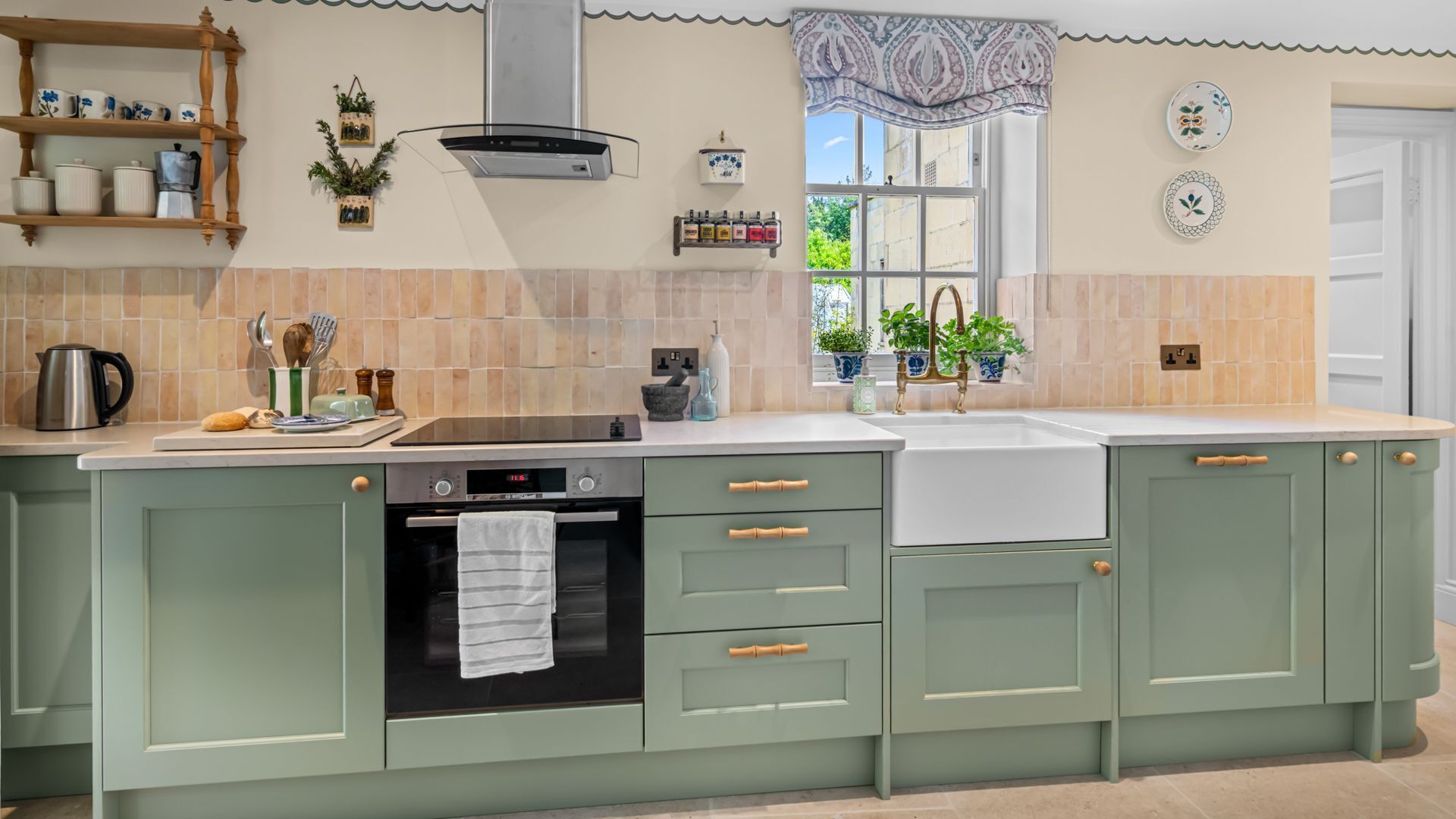 Una cocina con horno empotrado, placa de cocina y pileta. Hay armarios con pomos de madera y un ventanal con un frente decorado.