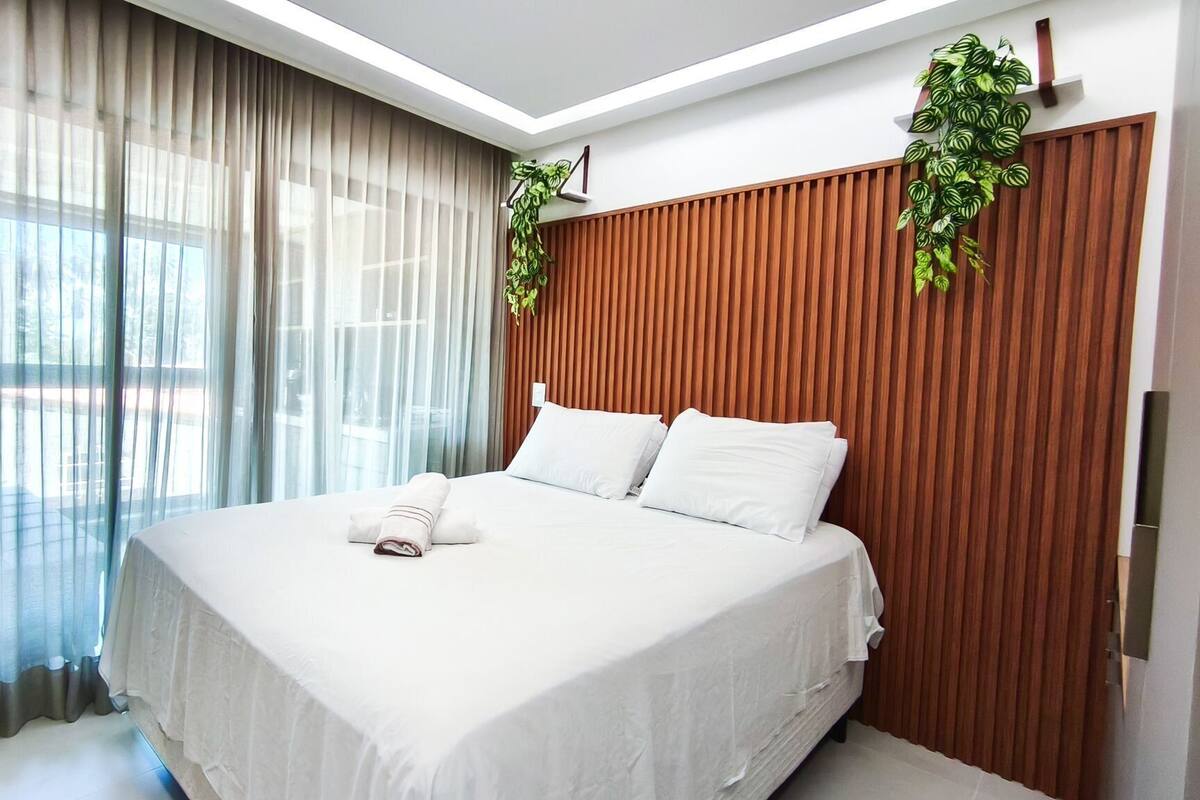 Ein modernes Hotelzimmer mit einem großen Bett, transparenten Vorhängen, einer Wand aus Holzpaneelen und einer Zimmerpflanze.