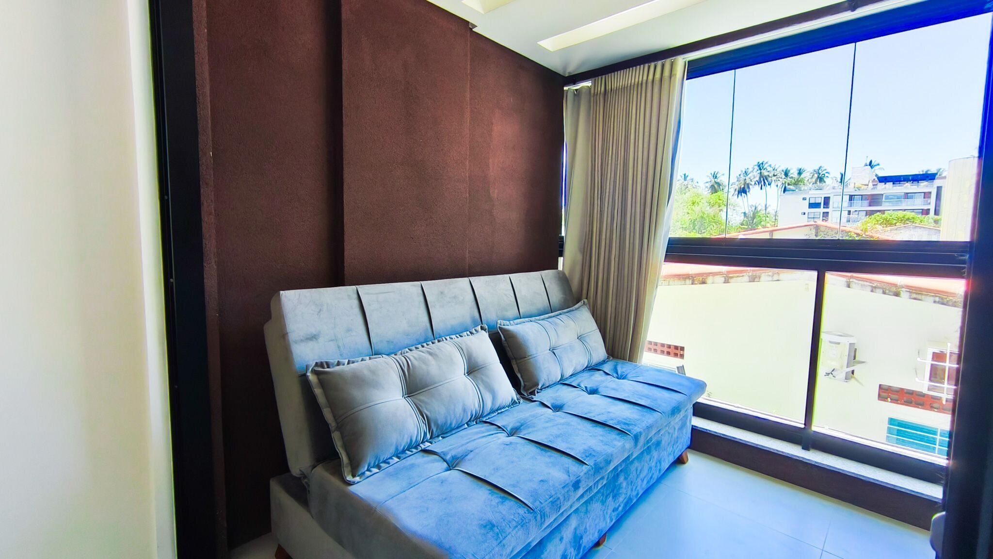 A room with a sofa against a wall, a large window with a view of buildings, and a curtain.