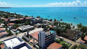 An aerial view of a coastal area with buildings, a clear blue sea, and a beach with palm trees.