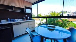 A modern kitchen with a microwave, sink, and cabinets, a round table with chairs, and a large window with a view of greenery and buildings. - Rooftop Tamandaré - Praia dos Carneiros (Tamandaré)