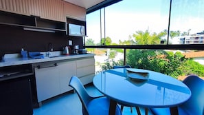 Een moderne keuken met een magnetron, spoelbak en kasten, een ronde tafel met stoelen en een groot raam met uitzicht op groen en gebouwen.