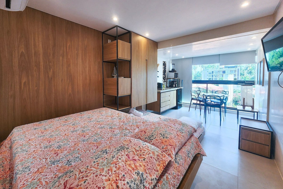 Quarto com cama, guarda-roupa de madeira, cozinha compacta e varanda com mesa e cadeiras.