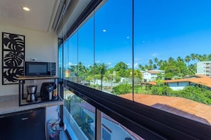 A kitchen with a microwave, coffee maker, and refrigerator, large windows with a view of palm trees and buildings. - Rooftop Tamandaré - Praia dos Carneiros (Tamandaré)