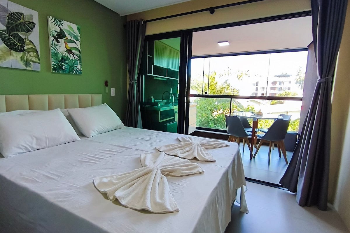 Quarto de hotel com cama, duas almofadas, toalha, parede verde e varanda com cadeiras.