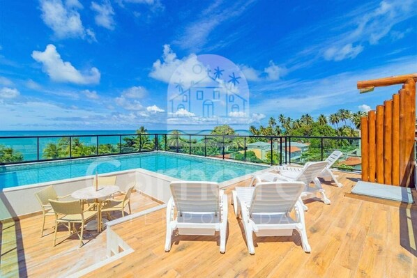 A rooftop pool area with lounge chairs and a table, overlooking a clear blue sky and a distant building.