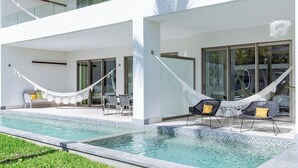 Un espace moderne aménagé au bord de la piscine, avec une façade blanche, des portes vitrées et deux hamacs.
