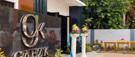 Entrada de hotel com uma placa de destaque, área de recepção com mesa e cadeiras, e um pequeno jardim com plantas e um pilar decorativo.