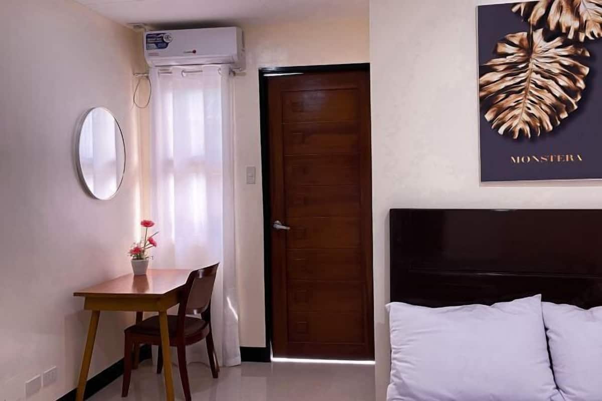 Habitación de hotel con cama, puerta de madera, mesa de comedor con sillas, espejo y aire acondicionado instalado en la pared.