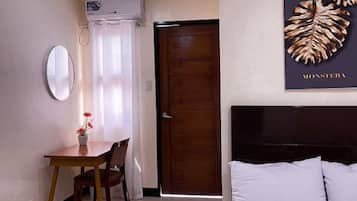 A hotel room with a bed, a wooden door, a dining table with chairs, a mirror, and a wall-mounted air conditioner.