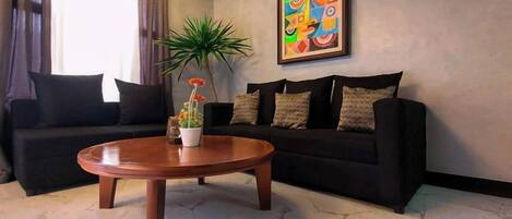 Una sala de estar con un sofá negro, una mesa de centro redonda de madera y una planta en maceta.