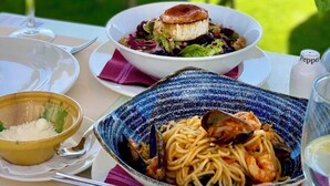 Una mesa dispuesta con ensalada, pasta y un postre, con vista al mar de fondo.