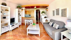 Sala de estar con sofá gris, mesa de centro blanca y estanterías con objetos decorativos. Se ve una zona de cocina al fondo con una mesa y sillas.