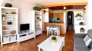 Un salón moderno con televisor, estanterías blancas, un comedor con mesa y sillas, y una cocina al fondo.