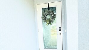Un couloir étroit avec une porte blanche ornée d’un panneau en verre dépoli et d’une guirlande. Un paillasson sur lequel est inscrit « SOYEZ NOS invités » est placé à l’entrée.