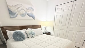 Un lit bien fait, avec une couette blanche et deux oreillers, une table de chevet avec une lampe, une œuvre d’art abstraite accrochée au mur et une porte ornée de panneaux décoratifs.