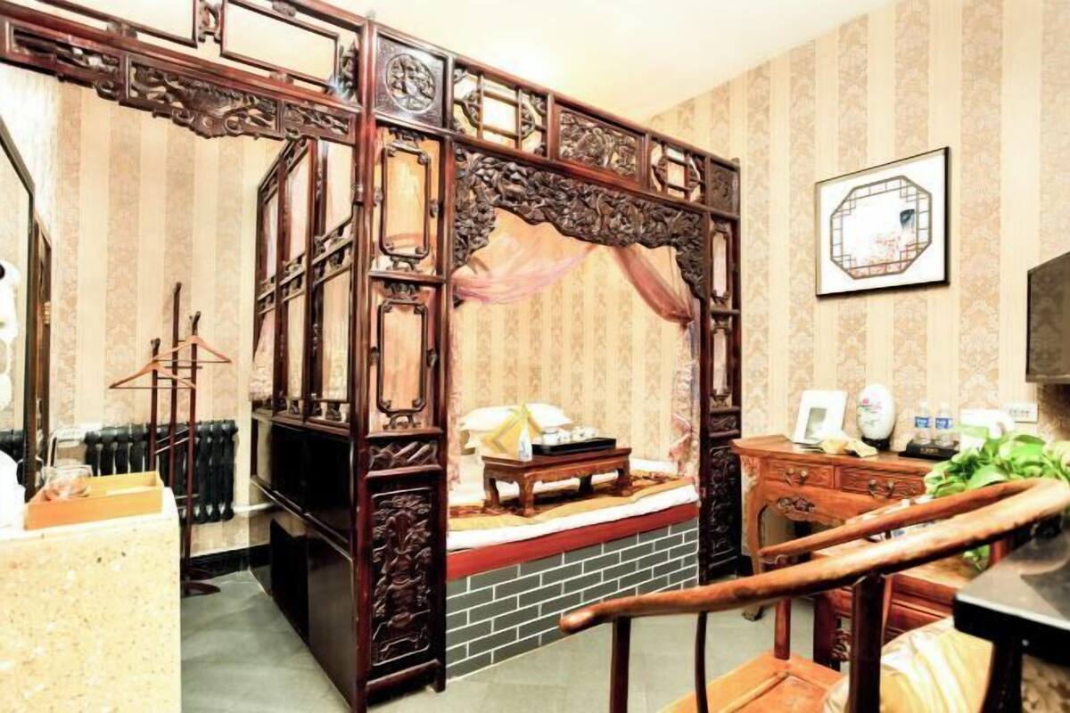 Una habitación tradicional con un separador de madera, una cama con cabecera, una mesita pequeña, una silla, un radiador y un cuadro en la pared.
