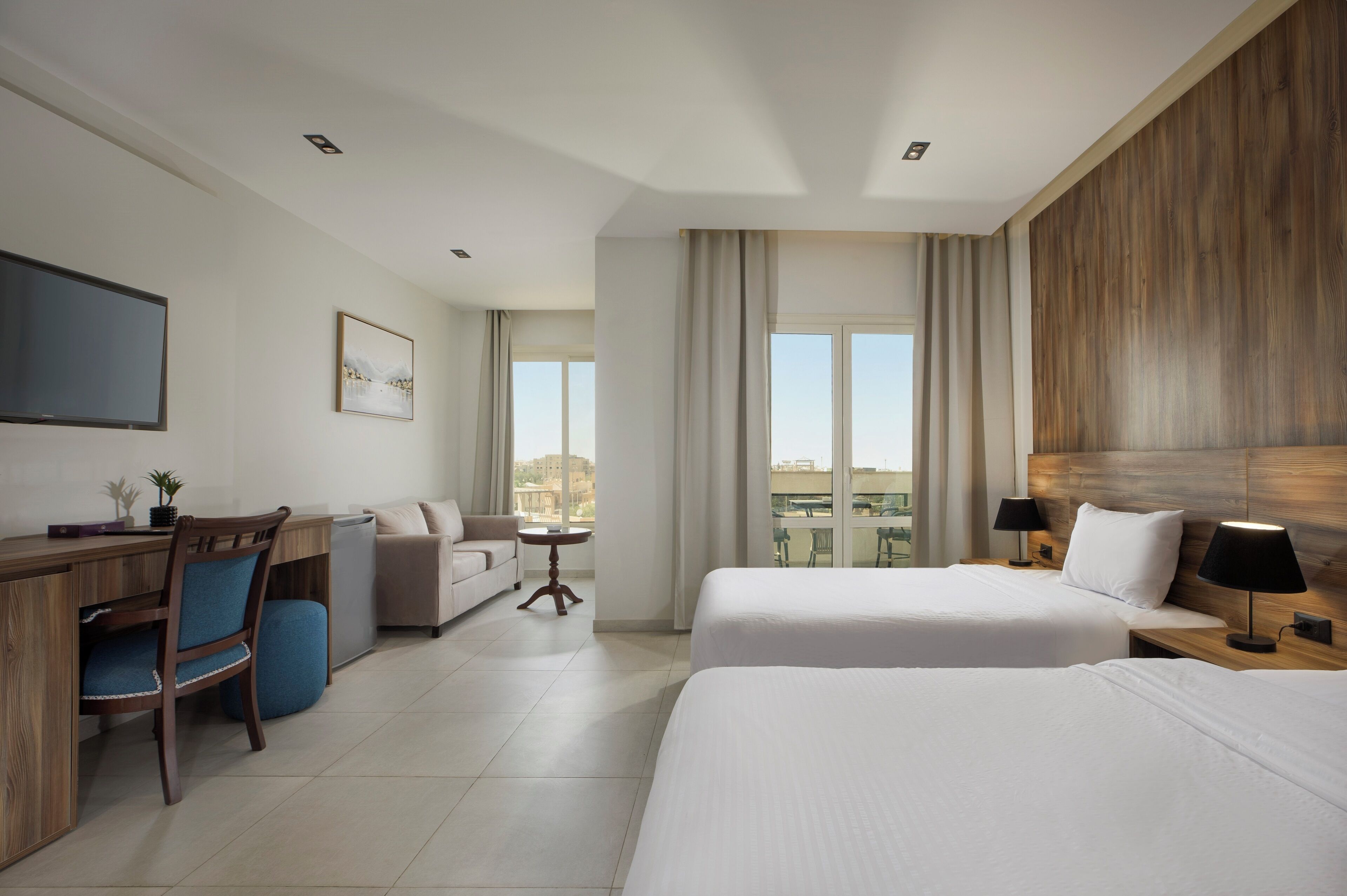 Ein Hotelzimmer mit zwei Betten, einem Schreibtisch, einem Sessel, einer Couch, einem Fernseher und Blick auf die Stadt.