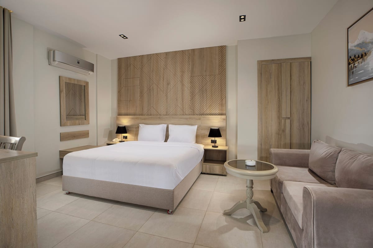 A modern hotel room with a large bed, bedside tables with lamps, a round table, a sofa, and a wooden headboard.