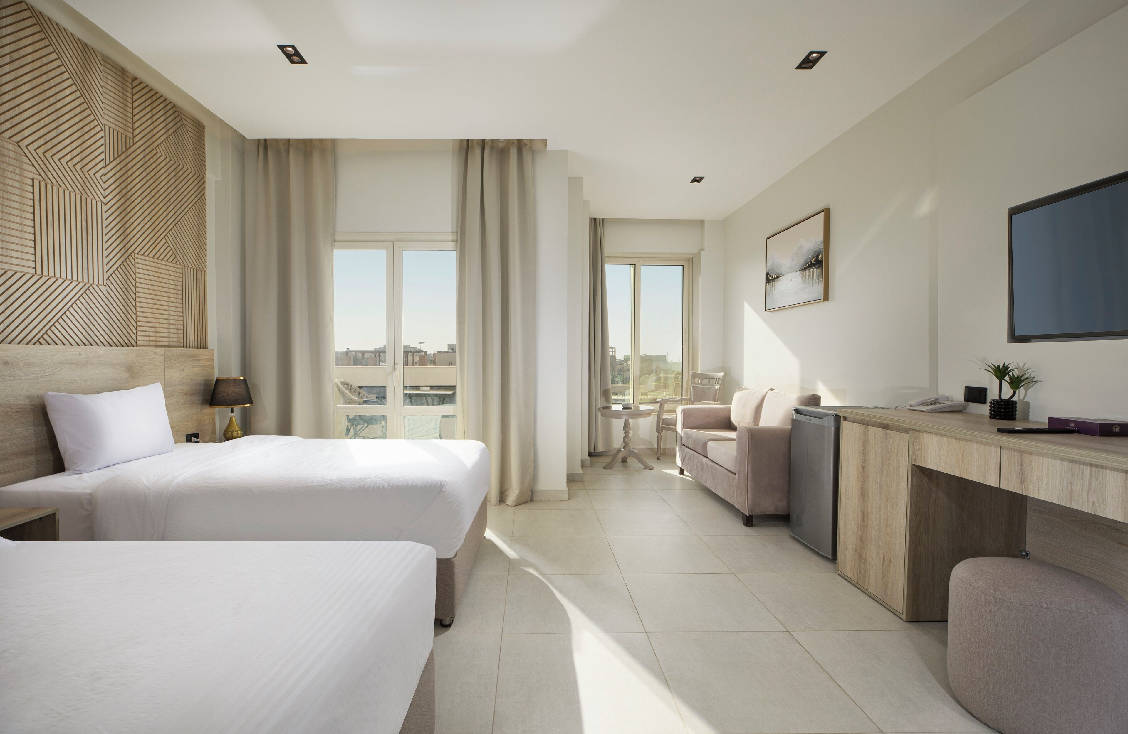 A modern hotel room with two beds, a desk, a chair, a small table, and a view of the city.