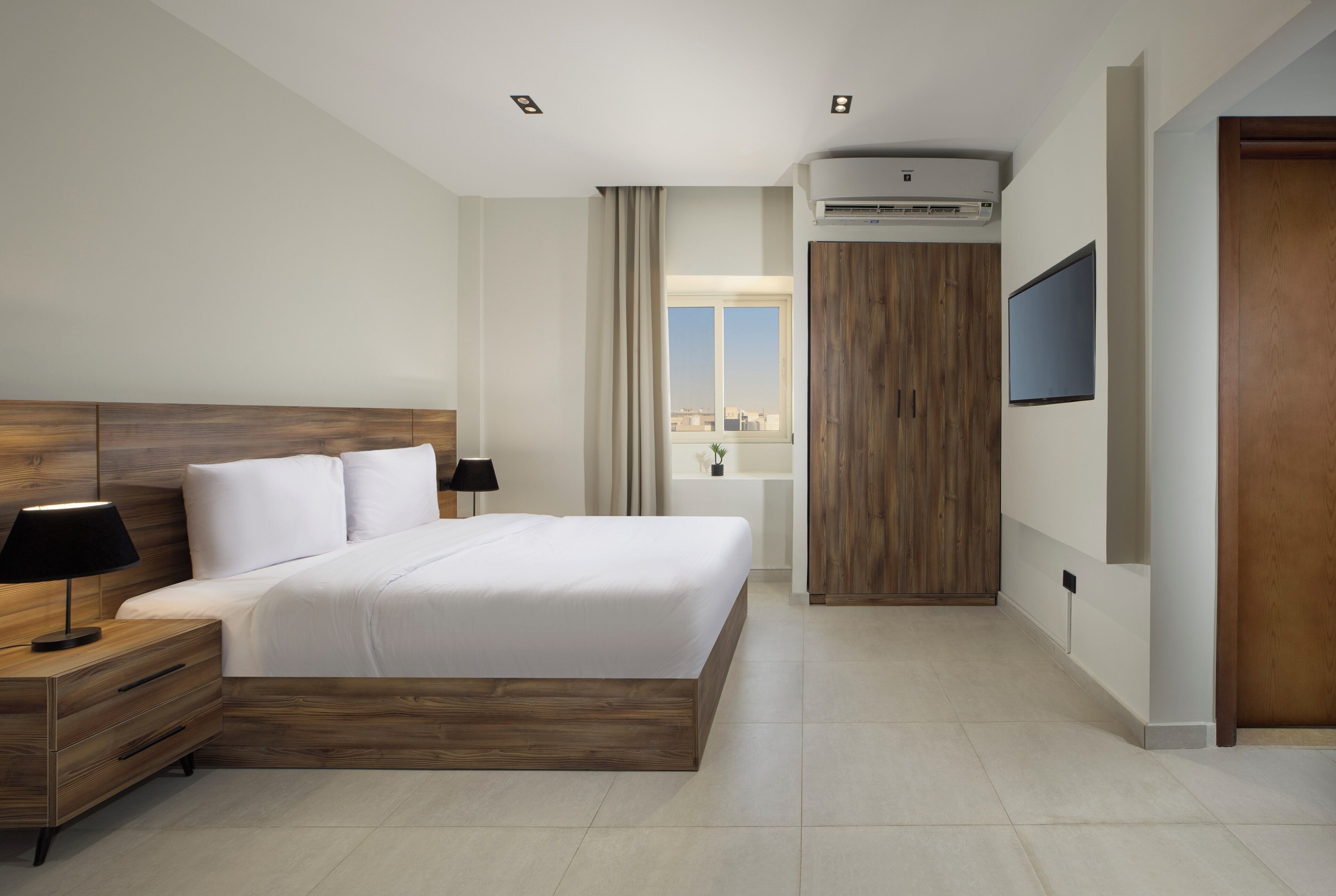 A modern hotel room with a wooden headboard bed, a nightstand with a lamp, a flat-screen TV mounted on the wall, and a large window offering a city view.
