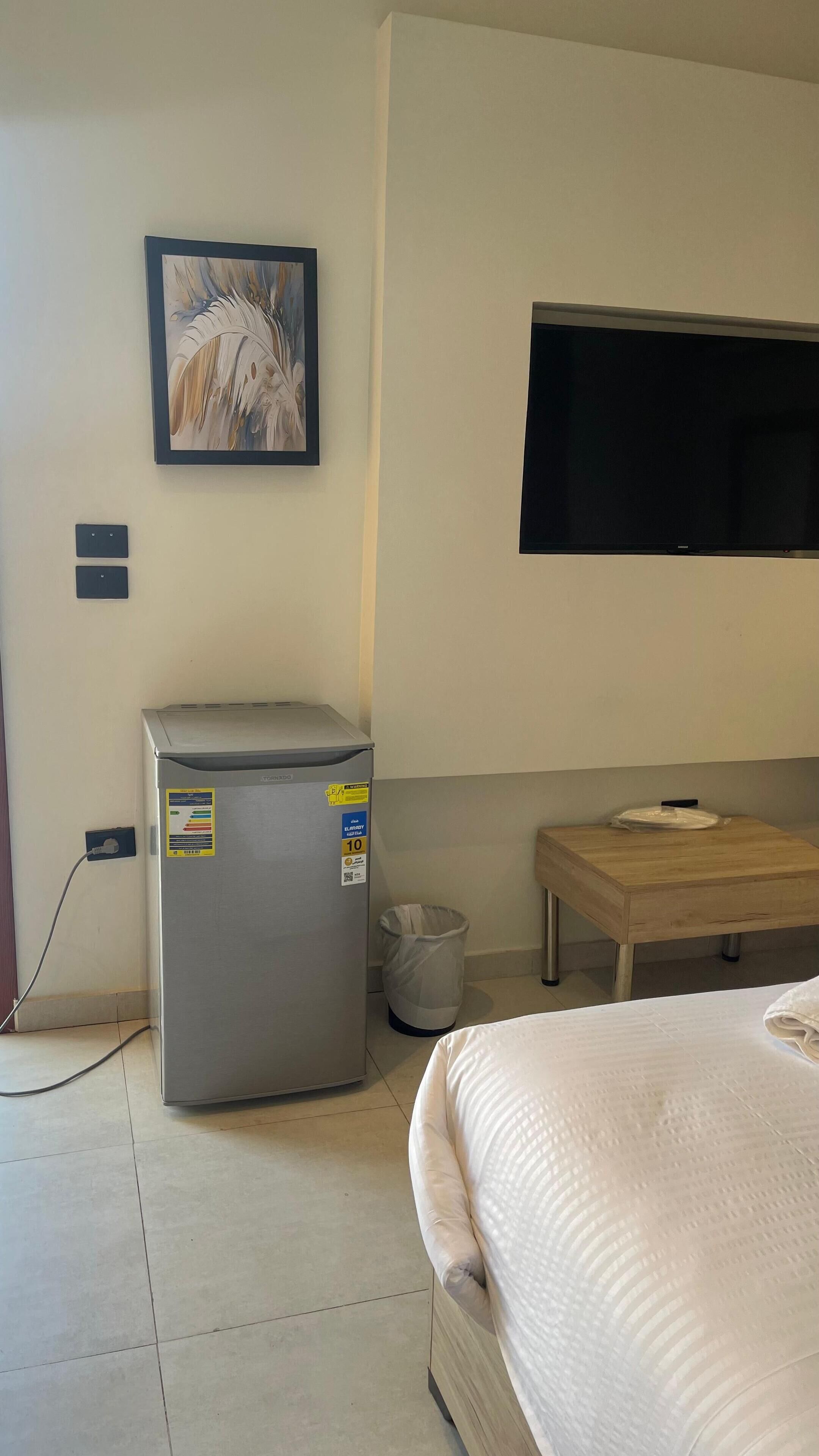Hotellihuone, jossa on sänky, pieni jääkaappi, roskakori, puinen pöytä, seinälle asennettu televisio ja taulu seinällä.