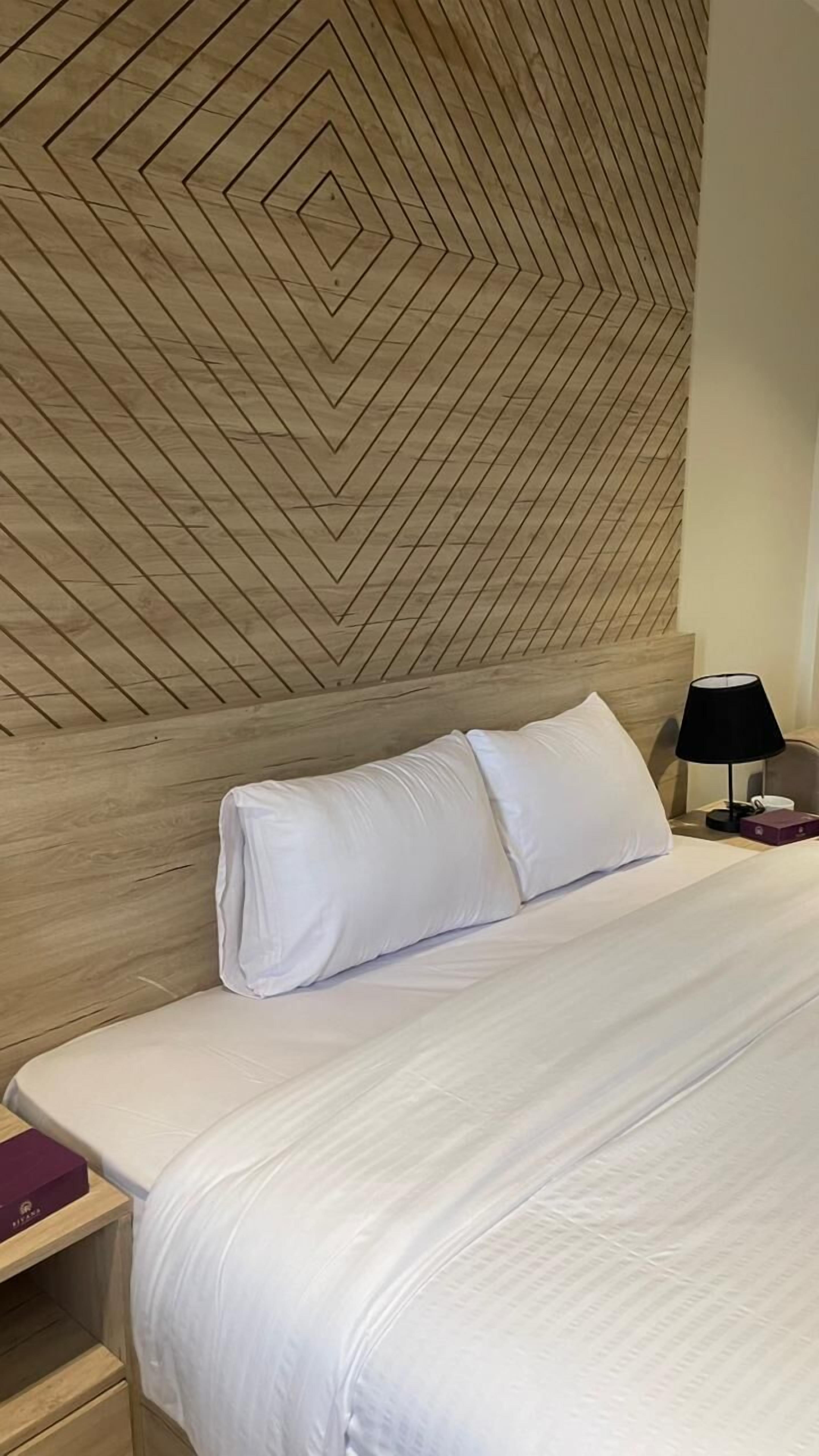 Una cama bien hecha con sábanas blancas, un cabecero de madera con un diseño geométrico, una mesita de noche con una lámpara y una caja morada sobre la mesita.