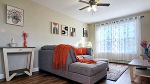 A living room with a grey sofa, a small table, a ceiling fan, and a window with sheer curtains.