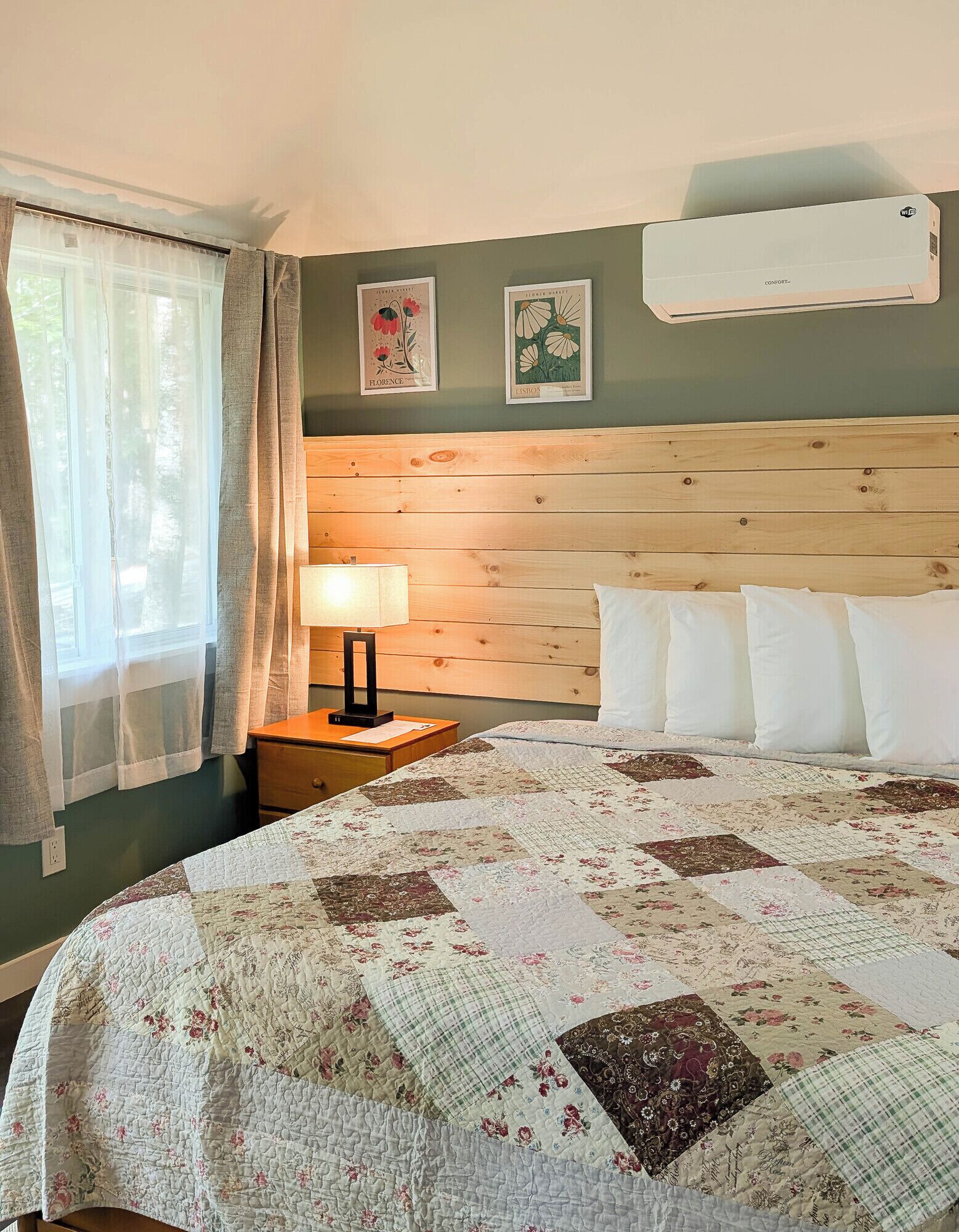 A hotel room with a wooden headboard, a quilted bedspread, a nightstand with a lamp, and two framed pictures on the wall.