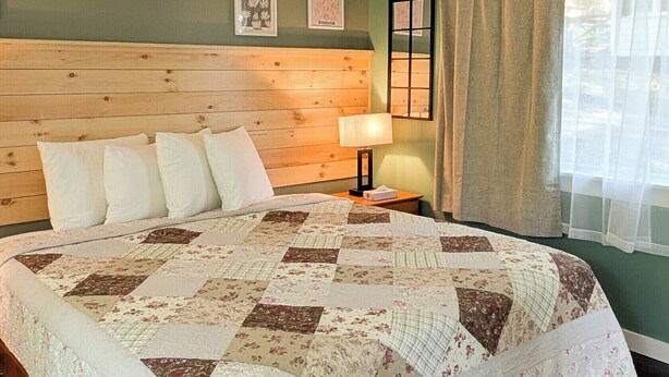 Un dormitorio con cabecera de madera, colcha acolchada, lámpara de noche y ventana con cortinas.