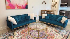 Una sala de estar moderna con dos sofás color turquesa, una mesa de centro redonda, un tapete colorido y una cocina al fondo.