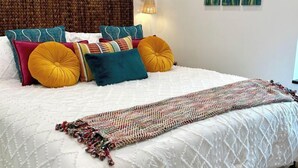 Una cama con colcha blanca, varios almohadones de colores y un cabecero tejido.