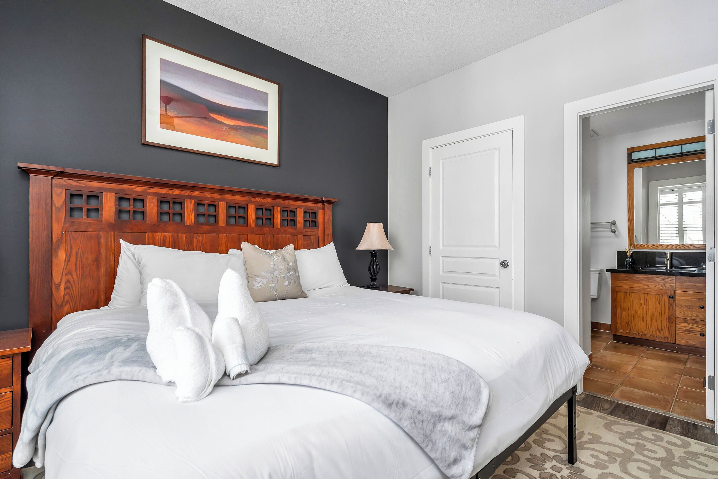 A neatly made bed with white linens and two white towels, a wooden headboard, a framed picture on the wall, and a view into the adjacent room.