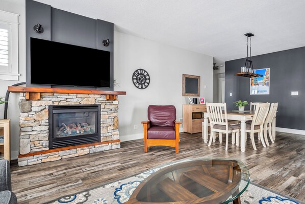 A modern living room with a stone fireplace, a flat-screen TV, a dining area with a table and chairs, and a decorative clock on the wall. - Rivergrass Condos (Blue Mountains)