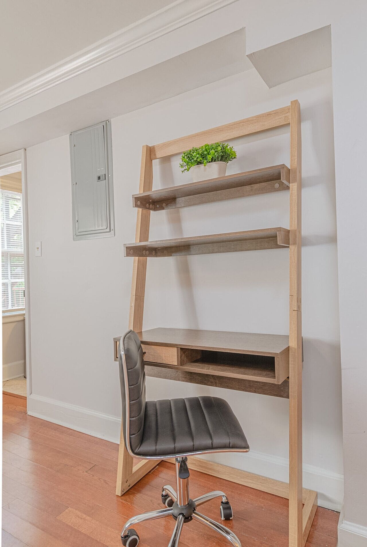 A corner desk with a chair, a potted plant on the top shelf, and a white wall with a vent.