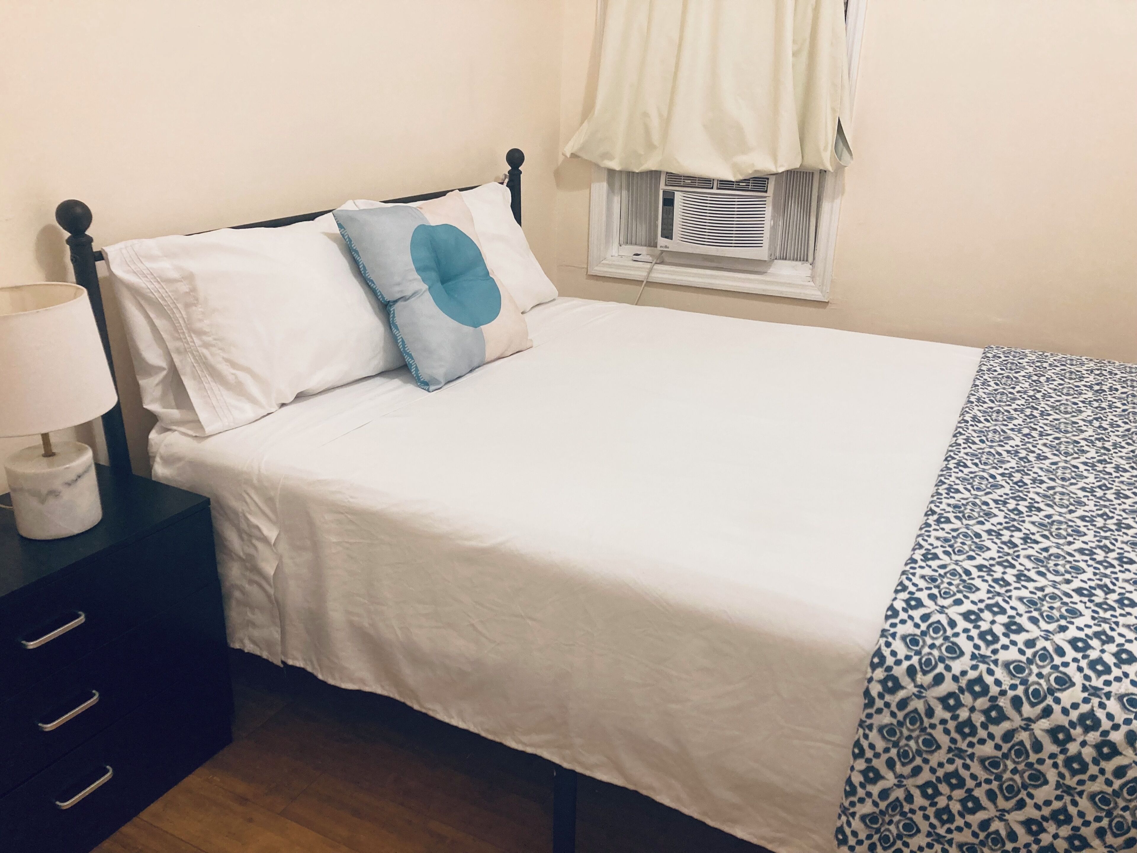 Una cama con ropa blanca, una almohada azul, un plaid estampado, una lámpara de noche y una ventana con aire acondicionado.