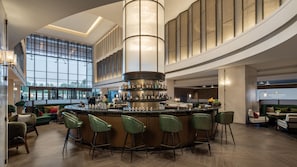 Un espace bar moderne doté d’un bar circulaire et de nombreux tabourets, de grandes fenêtres offrant une vue panoramique et d’un coin salon avec un canapé et des fauteuils verts.