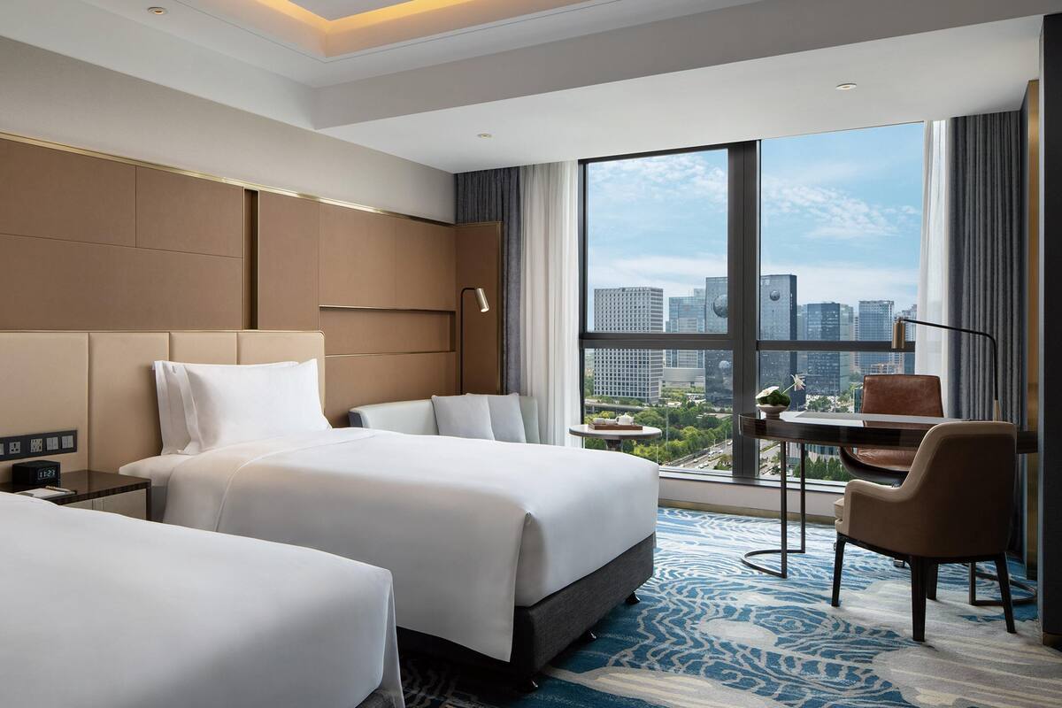 Habitación de hotel con dos camas, un escritorio, una silla y vistas a la ciudad, además de un ventanal grande.