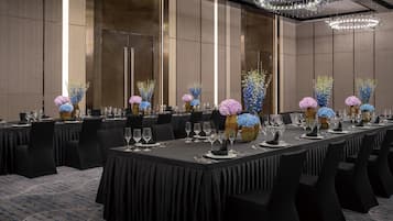 Un salón de conferencias con mesas largas dispuestas para un evento formal, con centros de mesa florales y una lámpara de araña moderna.