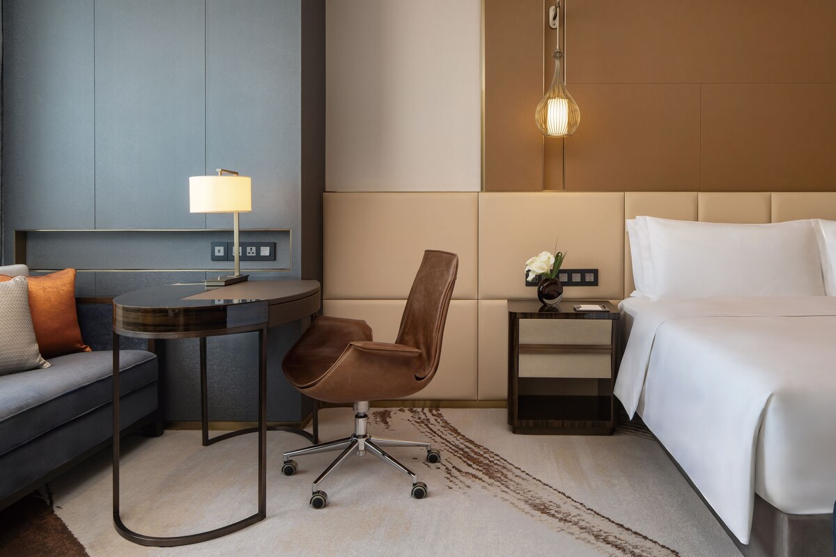 A hotel room with a bed, a desk, a chair, a lamp, and a nightstand with a vase of flowers.