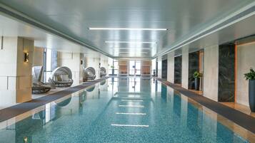 Una piscina interior larga con agua cristalina, rodeada de paredes con un diseño moderno y equipada con zonas de descanso a lo largo de los lados.