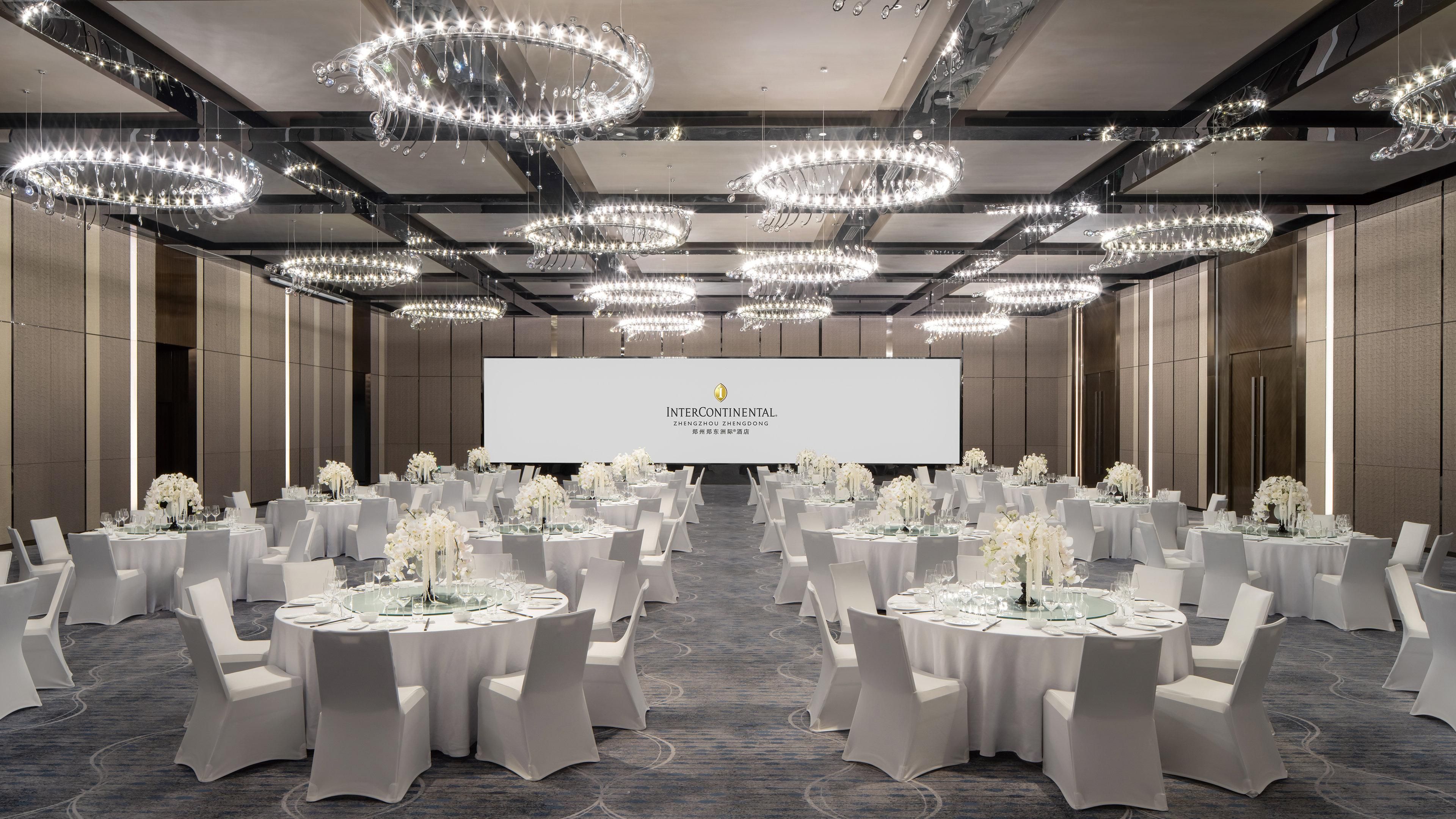 Een congreszaal met meerdere tafels gedekt voor een formeel evenement, met een groot scherm en elegante kroonluchters.