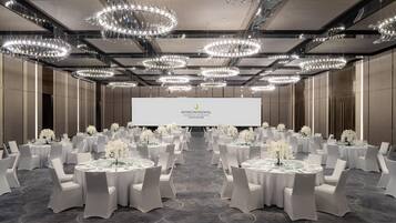 A conference room with multiple tables set for a formal event, featuring a large screen and elegant chandeliers.