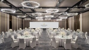 Een congreszaal met meerdere tafels gedekt voor een formeel evenement, met een groot scherm en elegante kroonluchters.