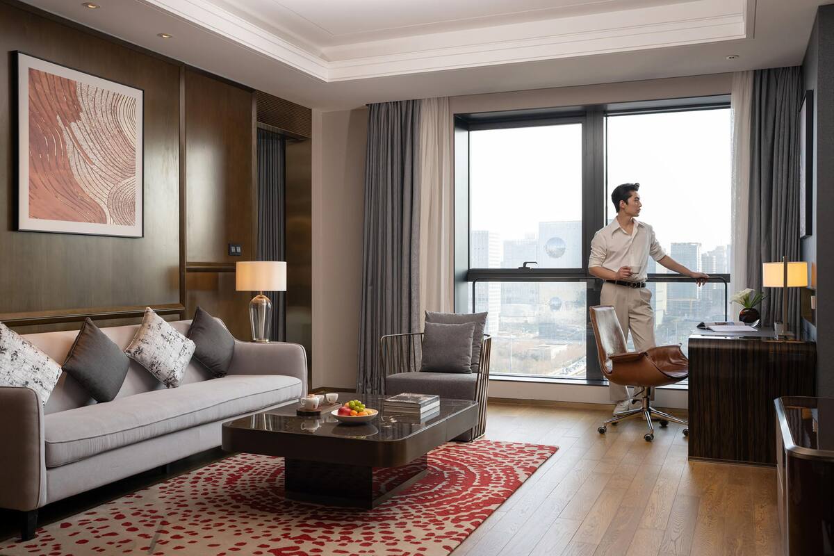 Habitación de hotel moderna con sofá, escritorio, silla, mesa baja con frutero y ventana grande con vista a la ciudad.